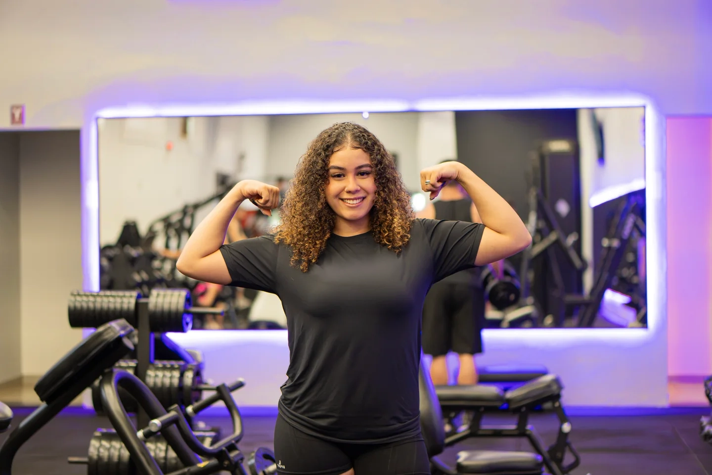 Isys Karoline sorrindo e flexionando os braços em uma academia